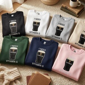K&ouml;nnte beinhalten: Eine Auswahl an Sweatshirts mit Rundhalsausschnitt in verschiedenen Farben, darunter Schwarz, Wei&szlig;, Grau, Beige, Gr&uuml;n, Marineblau und Rosa. Jedes Sweatshirt zeigt eine Grafik eines Guinness-Pints mit dem Text "I NEED VITAMIN G".