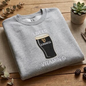 K&ouml;nnte beinhalten: Ein hellgraues Sweatshirt mit dem Text "I NEED VITAMIN G" und einer Guinness-Bierglas-Grafik. Das Sweatshirt liegt gefaltet auf einer Holzoberfl&auml;che, zusammen mit einer Sukkulente, einer Schere und anderen dekorativen Elementen.