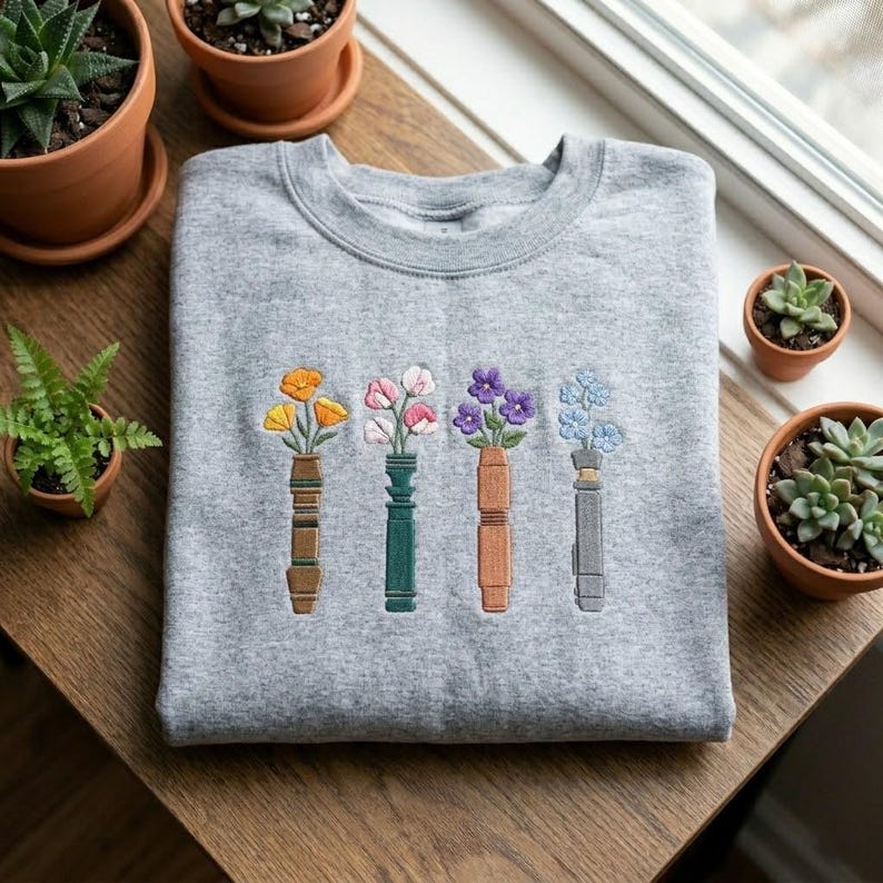 Puede incluir: Una sudadera gris jaspeada con un dise&ntilde;o floral bordado. El dise&ntilde;o muestra cuatro jarrones estilizados, cada uno con un tipo diferente de flor en tonos amarillos, rosas, morados y azules. Los jarrones son en tonos marrones, verdes y grises.