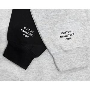Może przedstawiać: Zbliżenie dw&oacute;ch rękaw&oacute;w bluzy, jeden czarny i jeden szary. Oba rękawy mają biały nadruk "CUSTOM NAME/TEXT ICON". Rękawy wykonane są z miękkiego, wygodnego materiału.