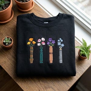 Puede incluir: Sudadera negra con un dise&ntilde;o floral bordado. El dise&ntilde;o incluye cuatro jarrones, cada uno con una flor diferente en tonos amarillos, rosas, morados y azules. La sudadera est&aacute; sobre una superficie de madera.