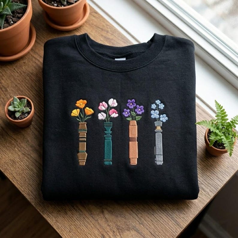 Puede incluir: Sudadera negra con un dise&ntilde;o floral bordado. El dise&ntilde;o incluye cuatro jarrones, cada uno con una flor diferente en tonos amarillos, rosas, morados y azules. La sudadera est&aacute; sobre una superficie de madera.