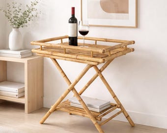 May include: A light brown bamboo bar cart featuring a rectangular top tray with a raised railing and a lower shelf. The cart has an X-shaped leg structure. A dark glass wine bottle and a clear glass filled with red wine sit on the top tray.