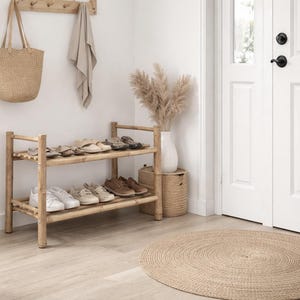 May include: A light wood shoe rack with two shelves holds various shoes. A woven tote bag hangs on the wall above the rack, next to a towel. A round woven rug sits on the floor, and a white vase with dried plants is on a woven basket.
