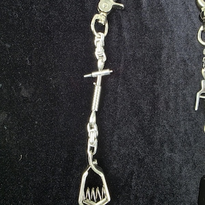 May include: A silver-toned metal accessory featuring a lobster claw clasp at the top, connected to a short chain segment, a cylindrical spring-loaded component, and a bottom clip with a serrated jaw mechanism designed for gripping items securely.