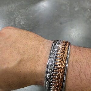 Puede incluir: Una pulsera de brazalete ancha con un diseño de múltiples hilos que presenta filas alternas de eslabones metálicos en tonos plateados y cobrizos. La pulsera tiene un borde de metal liso y sólido en la parte superior e inferior, creando un aspecto en capas.