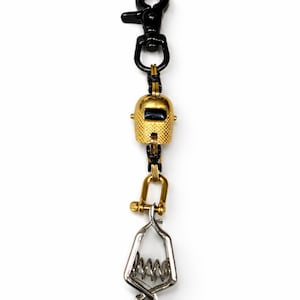 May include: A gold-colored welding helmet charm with a black clasp and a silver-colored clamp. The charm is suspended from a black metal chain. The welding helmet has a textured surface and a dark rectangular opening.