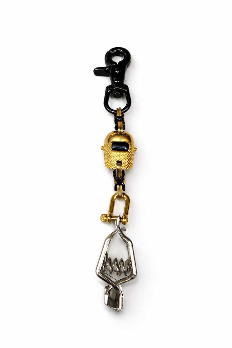 May include: A gold-colored welding helmet charm with a black clasp and a silver-colored clamp. The charm is suspended from a black metal chain. The welding helmet has a textured surface and a dark rectangular opening.