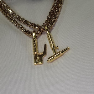 Op de afbeelding: Een goudkleurige Byzantijnse ketting met twee verschillende hangers. De ene hanger heeft de vorm van een hand die een gebaar maakt, en de andere heeft de vorm van een klein gereedschap of instrument. Beide hangers hebben een gepolijste afwerking.