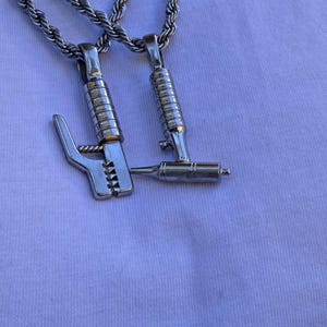 Op de afbeelding: Een zilverkleurige ketting met twee hangers in de vorm van lasgereedschap, een elektrodehouder en een lastoorts, hangend aan een gedraaide touwketting. De metalen hangers hebben een gepolijste afwerking en gedetailleerde texturen.