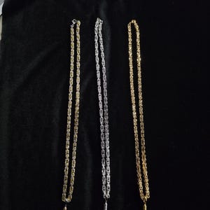 May include: Three metal necklaces featuring a thick, woven Byzantine chain design. Each necklace includes a small, detailed rifle-shaped pendant hanging from the bottom. Two of the necklaces are gold-toned, and one is silver-toned in colour.