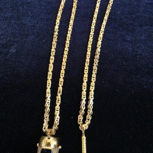 May include: Two gold-toned necklaces featuring thick, interlocking square-link chains. One necklace has a pendant shaped like a welding helmet with a dark rectangular visor, while the other features a pendant shaped like a welding torch tool.