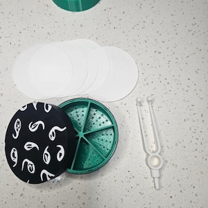 May include: An overhead shot shows a collection of items. There are several white circular objects, a green circular container, and a black circular lid with white abstract shapes. A white tuning fork-like tool is also present.