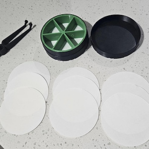 Pode incluir: Um recipiente circular de plástico preto com uma inserção verde de seis secções, uma tampa preta a condizer, uma ferramenta de plástico preto com duas pontas e nove discos de papel circulares brancos dispostos em três pilhas de três.