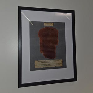Puede incluir: Obra de arte enmarcada que presenta un fragmento de texto antiguo, probablemente un pasaje b&iacute;blico. El texto es de color marr&oacute;n oscuro, sobre un fondo gris, y est&aacute; enmarcado en negro. El marco incluye un paspart&uacute; blanco, realzando la presentaci&oacute;n de la obra.