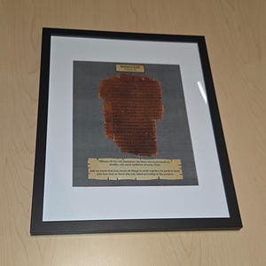 Puede incluir: Obra de arte enmarcada que presenta un fragmento de pergamino envejecido con texto. El pergamino marr&oacute;n est&aacute; montado sobre un fondo gris dentro de un passe-partout blanco. El marco negro rodea la obra, que incluye texto en la parte superior e inferior.
