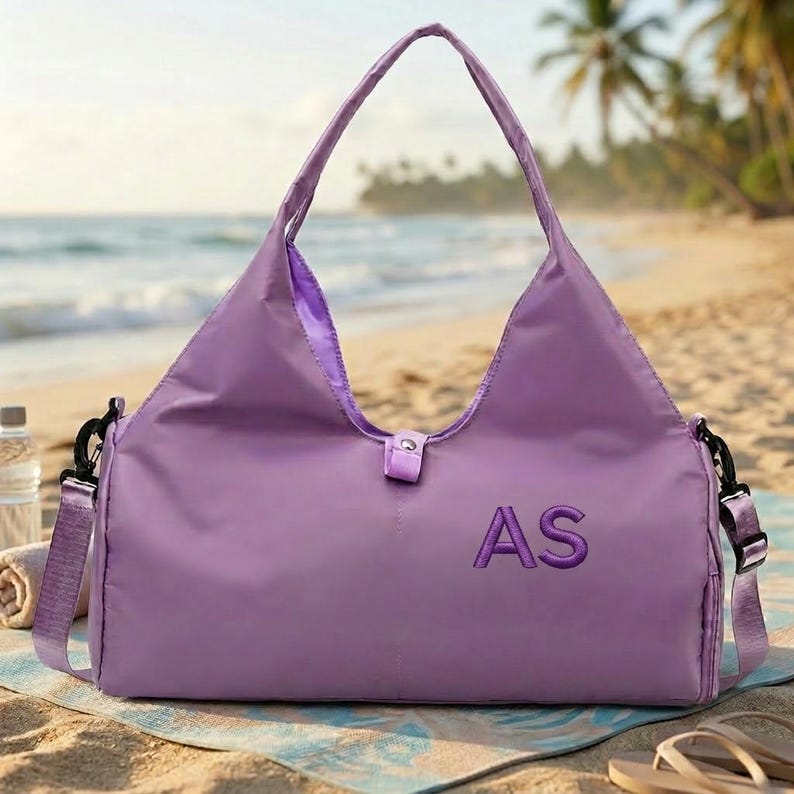 May include: A lavender tote bag with a shoulder strap and handles, resting on a beach towel. The bag features the embroidered initials "AS" in a darker purple thread. The background shows a beach with the ocean and palm trees.