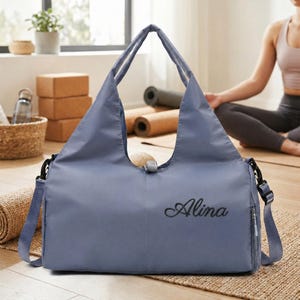 May include: A dusty blue yoga bag with the name "Alina" embroidered in black script. The bag has a shoulder strap and a top handle. The bag is sitting on a woven mat, with yoga mats and blocks in the background.