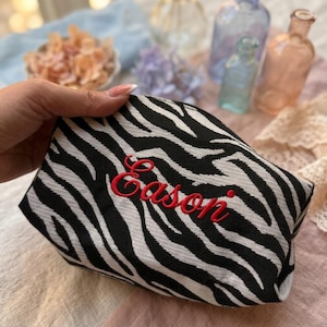 Puede incluir: Un neceser con estampado de cebra en blanco y negro con el nombre "Eason" bordado en rojo. El neceser está sujeto con una mano. Hay objetos decorativos en el fondo.