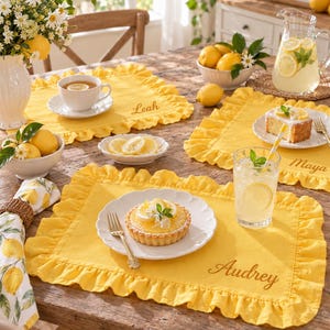 May include: A rectangular yellow fabric placemat featuring a ruffled border and the embroidered name Audrey in a cursive script. The placemat is set on a wooden table with a white plate holding a lemon tart, a glass of lemonade, and a fork.