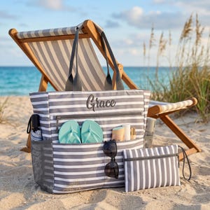 Könnte beinhalten: Eine grau-weiß gestreifte Strandtasche mit dem Namen "Grace" auf der Vorderseite. Die Tasche hat mehrere Taschen, darunter eine Seitentasche aus Netz, und enthält Flip-Flops, Sonnenbrille und Sonnencreme. Eine passende gestreifte Tasche ist ebenfalls enthalten.