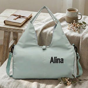 May include: A light green tote bag with the name "Alina" in black lettering. The bag has two handles and a detachable shoulder strap. A notebook, a mug of coffee, and flowers are in the background.
