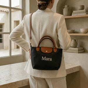 May include: Black handbag with brown leather accents, featuring a top handle and a shoulder strap. The bag has the name "Mara" embroidered in white. The bag is carried by a person wearing a cream-colored suit.