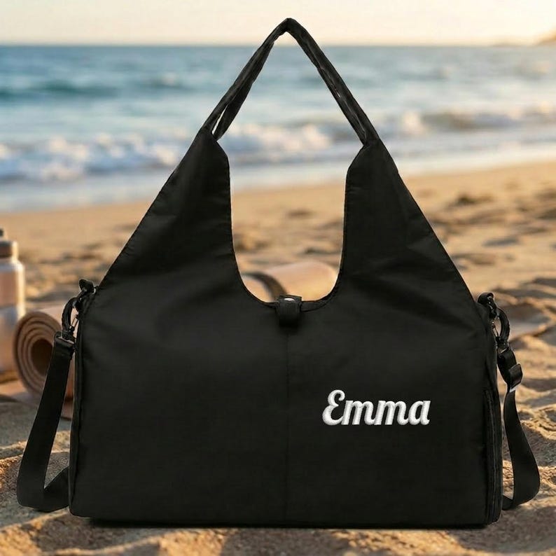 May include: Black tote bag with a shoulder strap, set on a sandy beach with the ocean in the background. The name "Emma" is embroidered in white script on the front of the bag. A water bottle and rolled yoga mat are visible.
