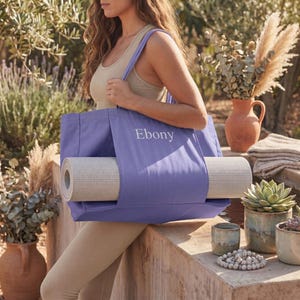 Puede incluir: Una bolsa de yoga lila con el nombre "Ebony" impreso en la parte delantera. La bolsa contiene una esterilla de yoga enrollada. La bolsa se lleva al aire libre.
