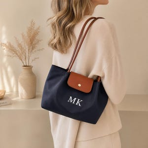 Pode incluir: Bolsa tote azul marinho com detalhes em couro castanho. A bolsa apresenta um fecho de aba castanho com um botão dourado e alças de couro castanho. As iniciais brancas "MK" são impressas na frente.