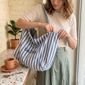 Puede incluir: Un bolso tote a rayas azules y blancas con una correa a juego. El bolso tiene un bolsillo frontal con el texto bordado "Est. 2026". El bolso está hecho de un material suave y lo sostiene una persona.