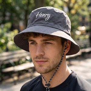 May include: Gray bucket hat with the name "Henry" in white script. The hat has a wide brim and a chin strap with a black and white cord. The hat is made of a lightweight material, suitable for outdoor wear.