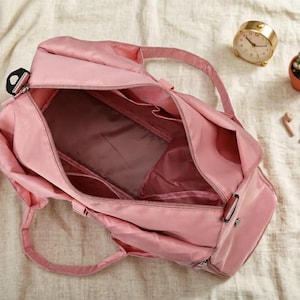 May include: A pink duffel bag with multiple compartments, open to reveal the interior. The bag has two handles and a shoulder strap. A gold clock, small potted succulent, and wireless earbuds are nearby.