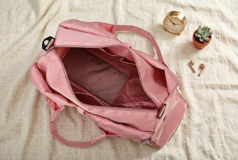 May include: A pink duffel bag with multiple compartments, open to reveal the interior. The bag has two handles and a shoulder strap. A gold clock, small potted succulent, and wireless earbuds are nearby.