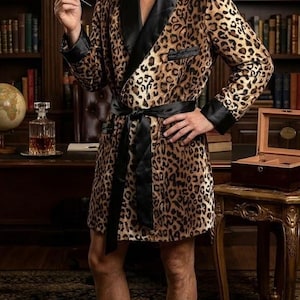 May include: A leopard print dressing gown with black satin trim and a matching belt. The gown is knee-length and worn with black loafers. The background features a library with bookshelves and a desk.