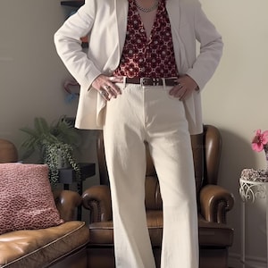 May include: A cream-coloured suit jacket and flared trousers paired with a burgundy floral print shirt and brown belt. The outfit is completed with brown shoes and a silver chain necklace. The look is reminiscent of 1970s fashion.