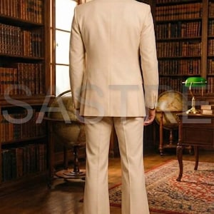 May include: A beige suit with a jacket and flared pants. The suit is worn by a person standing in a library with bookshelves and a globe. The suit has a classic, tailored look.