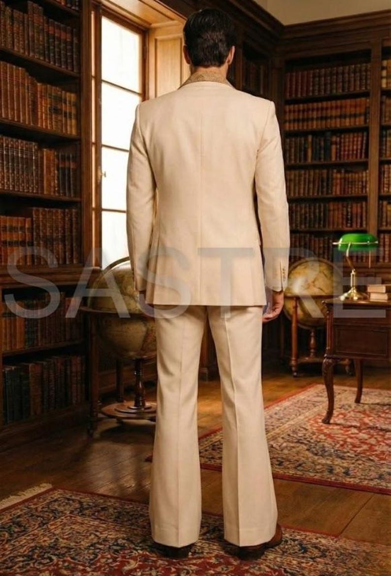 May include: A beige suit with a jacket and flared pants. The suit is worn by a person standing in a library with bookshelves and a globe. The suit has a classic, tailored look.