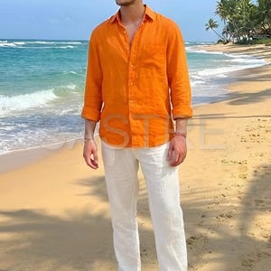 May include: An orange button-down shirt and white linen pants are worn on a beach. The shirt has long sleeves and is paired with white pants and brown sandals. The person is wearing sunglasses. The background includes the ocean, sand, and palm trees.