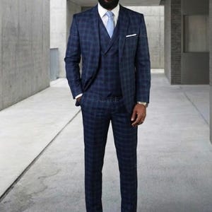 May include: A three-piece navy blue suit with a plaid pattern. The suit includes a jacket, vest, and trousers. The jacket has a notched lapel and a pocket square. The outfit is completed with a light blue tie and dark blue shoes.