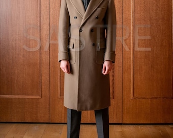 May include: A long, double-breasted, tan overcoat with a notched lapel and two rows of buttons. The coat has two front pockets and is worn with dark gray trousers and black dress shoes. The background is a wooden door.