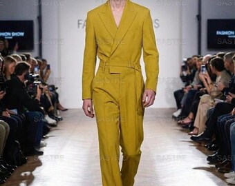 May include: A mustard yellow suit with a double-breasted jacket and matching trousers. The suit features a belt detail at the waist and is worn by a model on a runway. The background includes the text "FASHION SHOW" and the brand name "SASTRE".