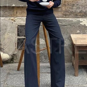 May include: Navy blue pinstripe trousers with a wide leg. The trousers are paired with a navy blue sweater. The person is holding a white teacup and saucer. The person is sitting on a wooden stool. The trousers are made of a lightweight fabric.