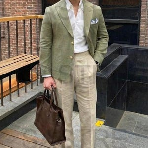 May include: A man in a green blazer, white shirt, and beige trousers, carrying a brown leather tote bag. He wears brown suede loafers. The setting includes a brick wall and a wooden deck.