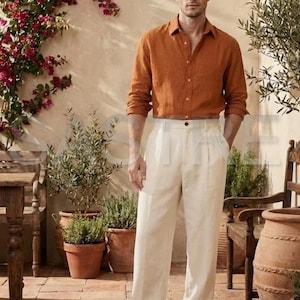 May include: A man wearing an orange long-sleeve button-down shirt and white pleated trousers. The outfit is paired with brown leather shoes. The setting is an outdoor patio with terracotta pots and flowering plants.