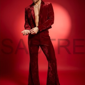 May include: A burgundy velvet suit, featuring a cropped blazer and flared pants. The outfit is worn with a silver chain and sunglasses. The background is a solid red color. The word "Satre" is partially visible in the background.