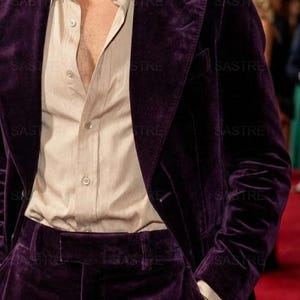 May include: A deep purple velvet suit, consisting of a blazer and trousers, paired with a light beige button-down shirt. The blazer has a classic two-button closure. The outfit is a formal ensemble, suitable for special occasions.