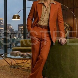 May include: A rust-colored suit with a matching jacket and wide-leg pants. The suit is paired with a patterned button-down shirt and a brown belt. The man is wearing brown leather shoes. The setting is a well-lit room with a city view.