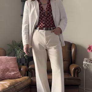 May include: A man in a white blazer, cream flared trousers, and a burgundy patterned shirt. He wears sunglasses and a gold chain. The outfit is reminiscent of 1970s fashion. The trousers are flared.