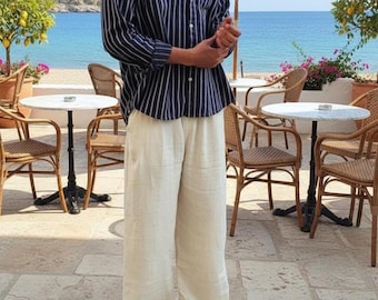 May include: A navy blue and white striped button-down shirt paired with cream-colored wide-leg pants. The person is wearing black sandals. The background includes a beach scene with tables, chairs, and umbrellas.